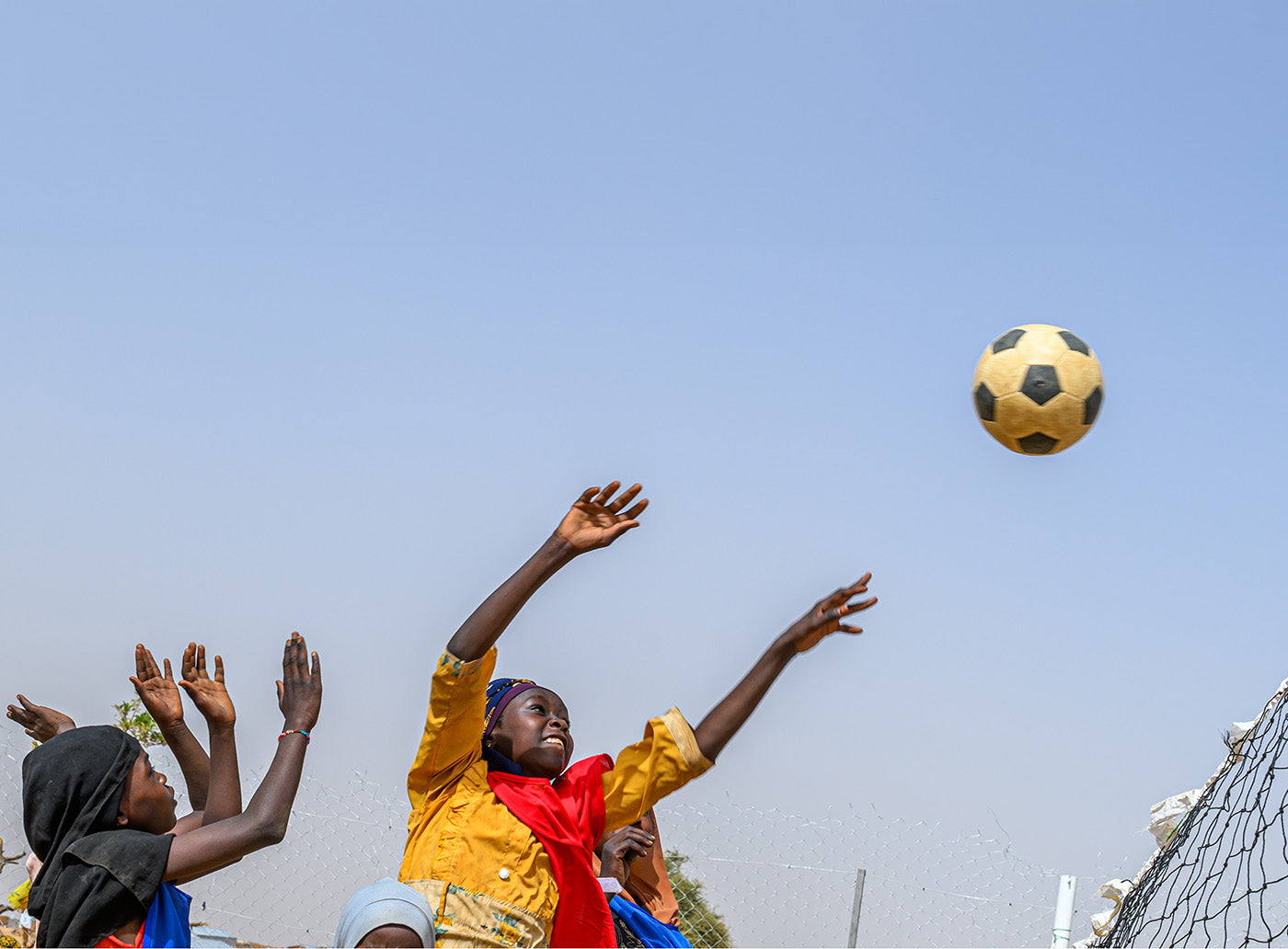 Un groupe d’enfants tendent les bras avec enthousiasme
vers un ballon de soccer en l’air près d’un but, sous un ciel bleu.