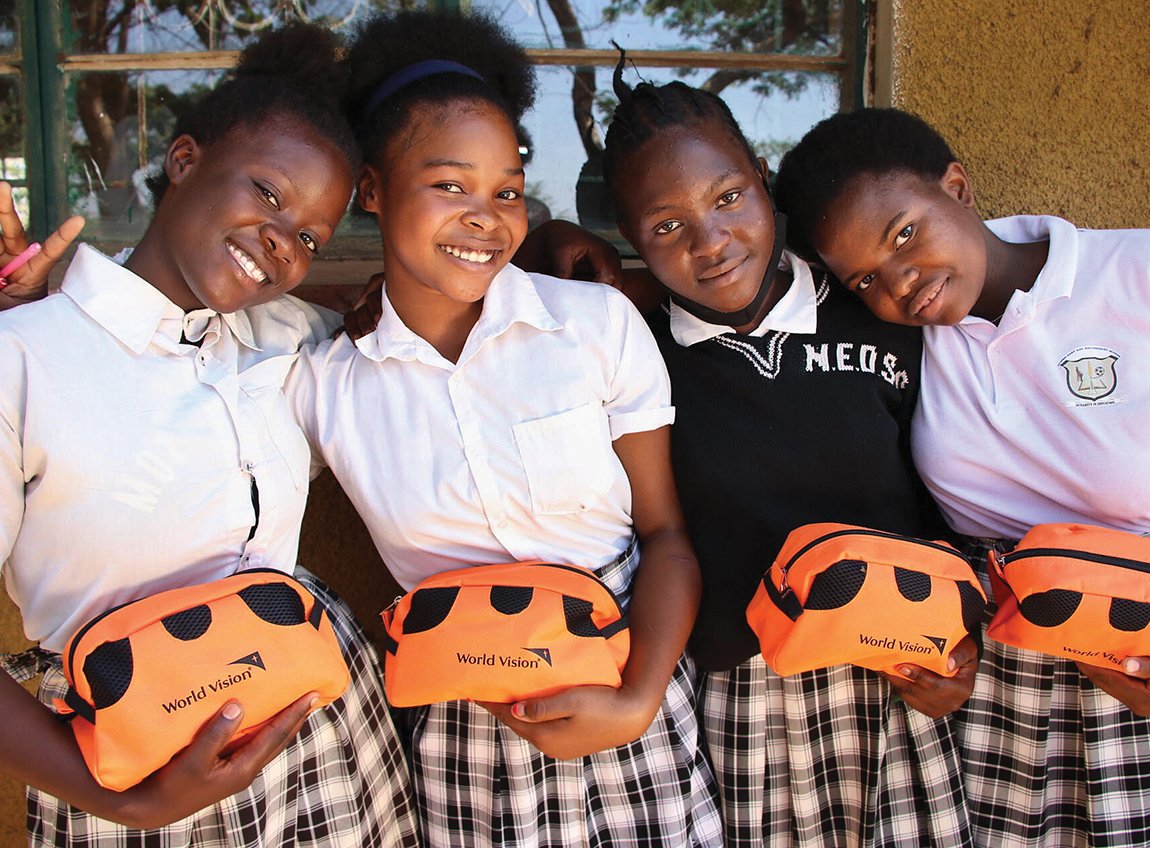 Quatre jeunes filles en uniforme avec des jupes à carreaux sourient vers l’objectif. Chacune tient une petite pochette de voyage qui contient une trousse d’hygiène.