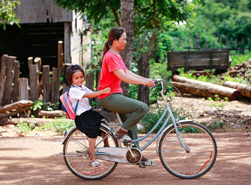 Une femme roule à vélo, avec un enfant souriant derrière elle. 
L’enfant porte un uniforme scolaire et un sac à dos. L’arrière-plan montre un paysage rural, vu arbres et des structures en bois.