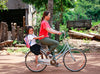 Une femme roule à vélo, avec un enfant souriant derrière elle. 
L’enfant porte un uniforme scolaire et un sac à dos. L’arrière-plan montre un paysage rural, vu arbres et des structures en bois.