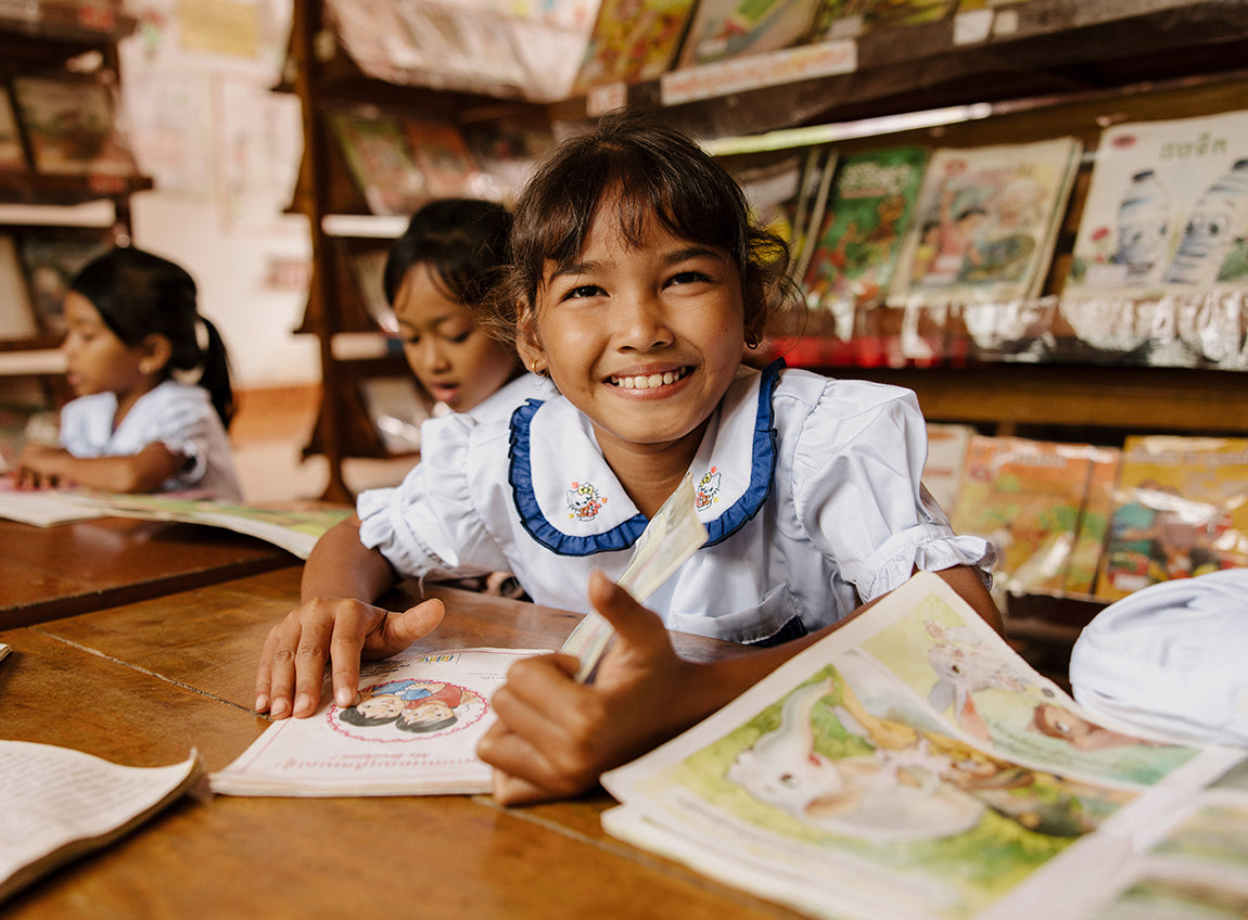 Une jeune fille dans une salle de classe sourit au photographe, un livre ouvert devant elle.