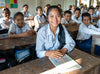 Une salle de classe d’élèves assis à des bureaux en bois, qui portent des uniformes et sourient, avec une élève au premier plan qui tient un livre.