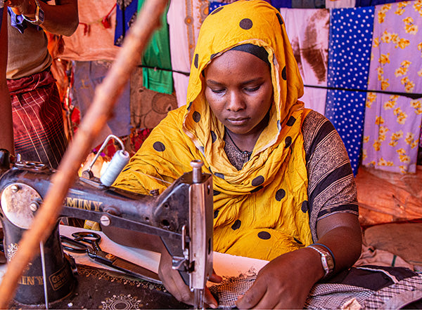 Une femme portant un foulard jaune travaille avec une machine à coudre, 
entourée de tissus colorés.