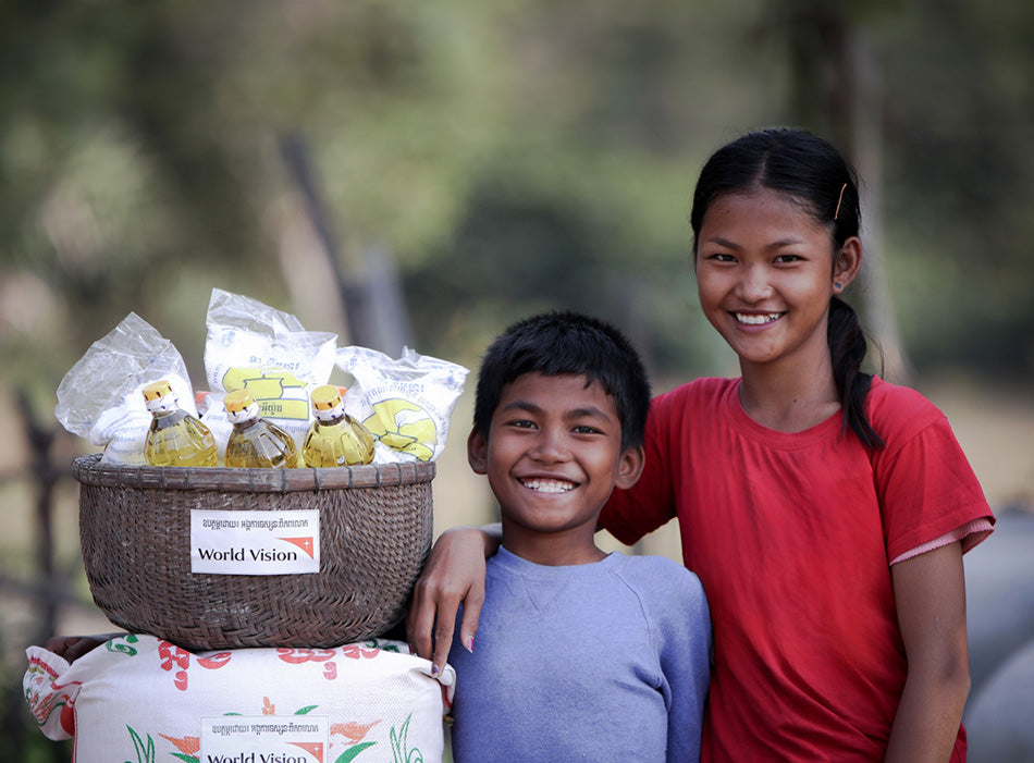 Une fille attrape son frère par les épaules et ils sourient tous les deux en se tenant près des articles alimentaires essentiels fournis par Vision Mondiale.