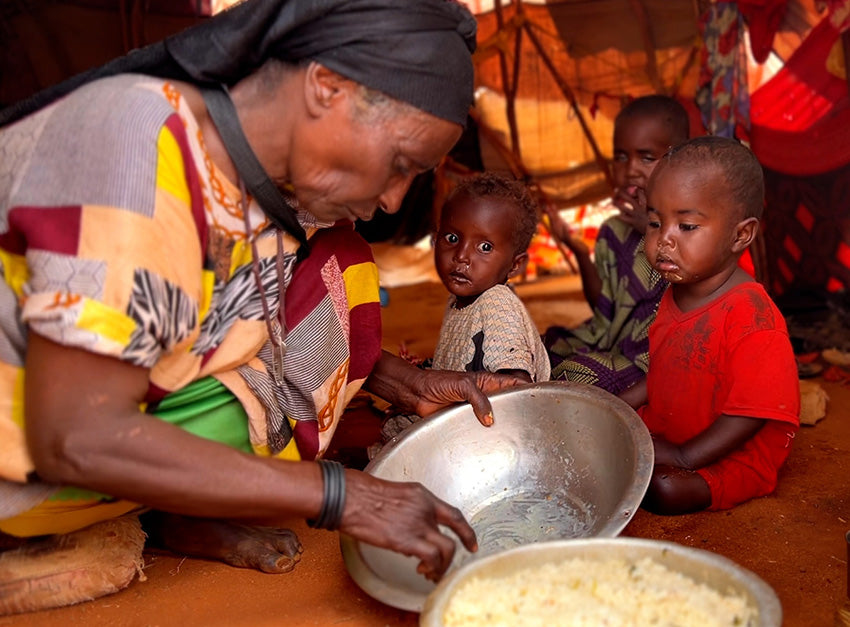 Une grand-mère sert à manger à partir d’un grand bol à ses trois petits-enfants assis à terre, dans une structure ressemblant à une tente.