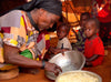 Une grand-mère sert à manger à partir d’un grand bol à ses trois petits-enfants assis à terre, dans une structure ressemblant à une tente.