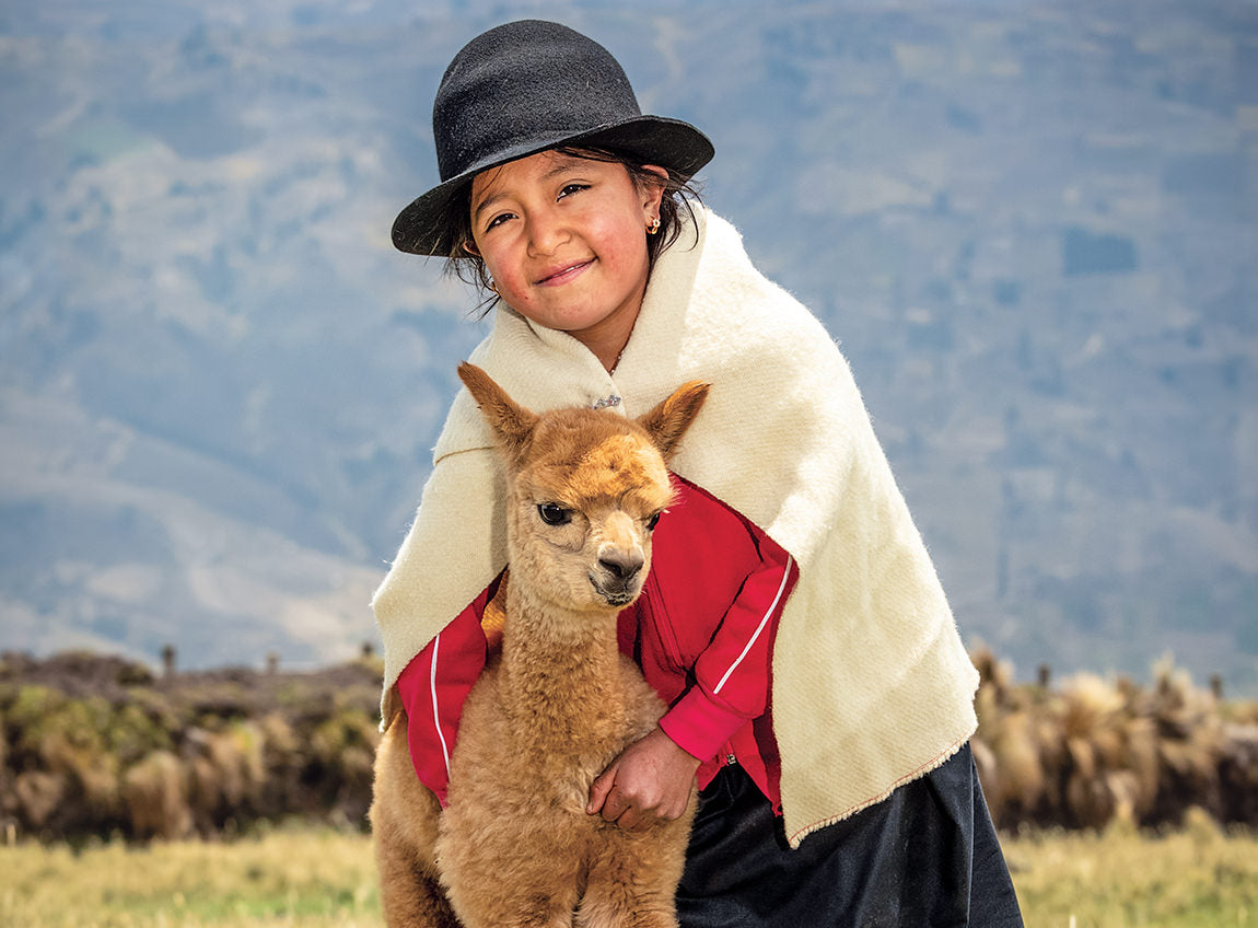 Dans un paysage en altitude, une jeune fille en tenue traditionnelle des Andes serre un alpaga dans ses bras.