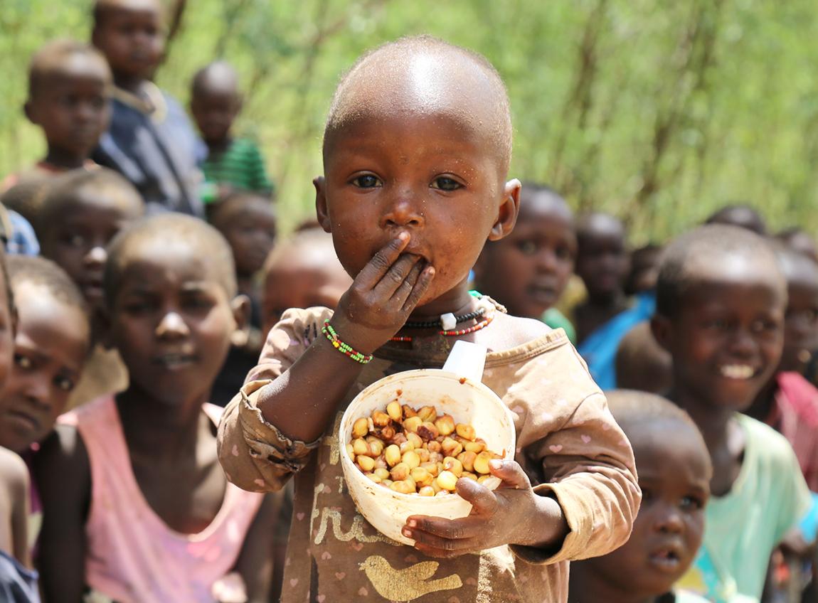Un très jeune enfant regarde vers l’objectif en mangeant à partir d’un contenant dans ses mains. Plusieurs autres enfants sont assis en arrière-plan.
