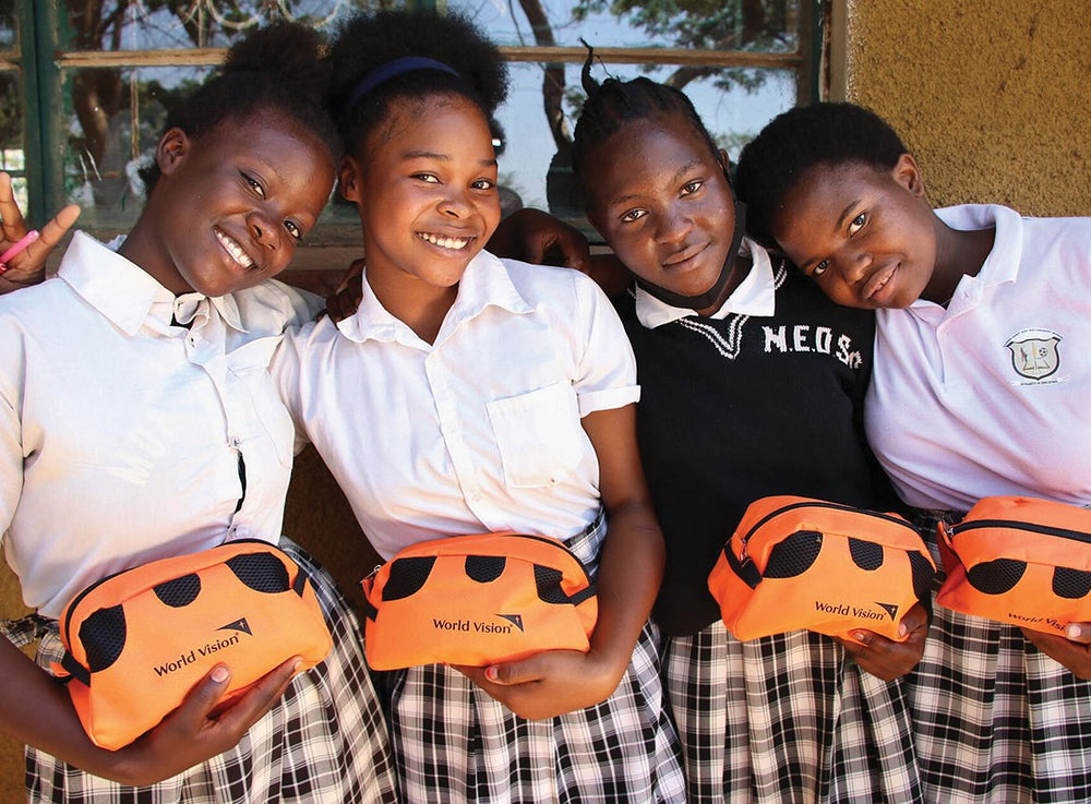 Quatre jeunes filles en uniforme avec des jupes à carreaux sourient vers l’objectif. Chacune tient une petite pochette de voyage qui contient une trousse d’hygiène.