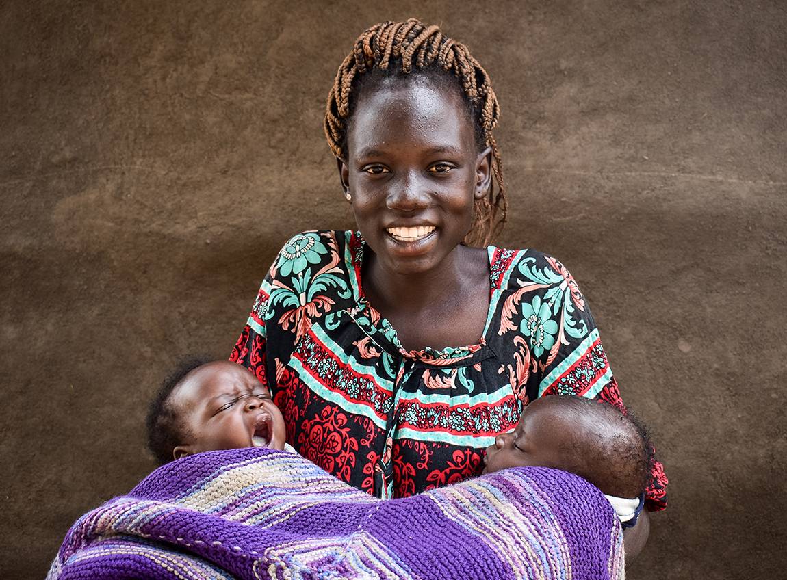 Une femme sourit vers l’objectif en tenant deux nouveau-nés enveloppés dans une couverture mauve.