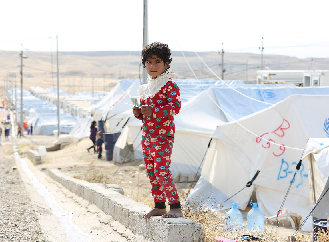 Une jeune fille en pyjama à fleurs se tient debout sur un mur de béton qui surplombe les tentes d’un camp de réfugiés.