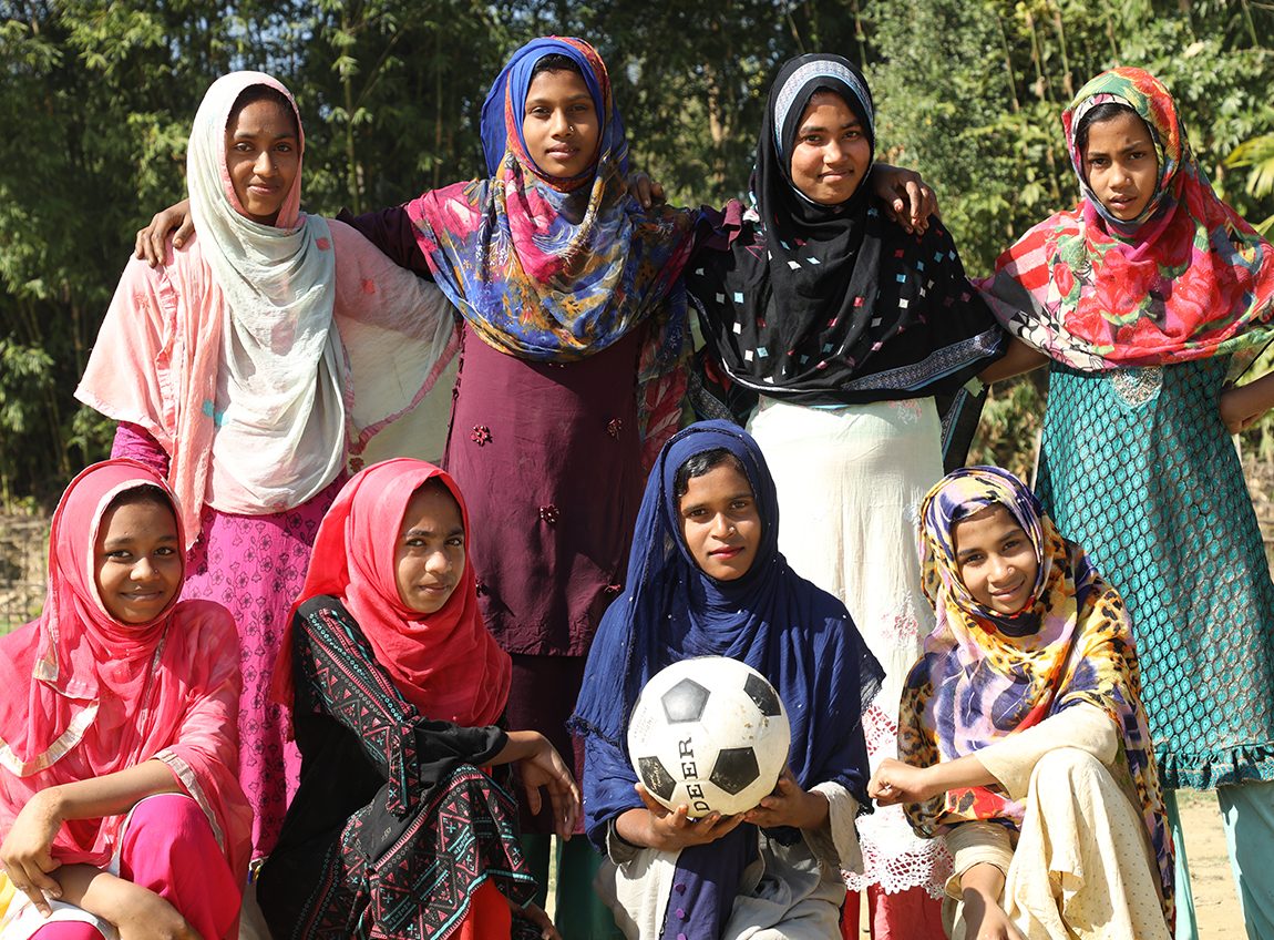 Huit jeunes filles en saris posent pour une photo. Une fille au milieu tient un grand ballon de soccer dans ses mains.