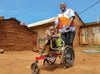 Un homme souriant portant un gilet World Vision orange et blanc pousse un enfant souriant dans un fauteuil roulant le long d’une route de terre.